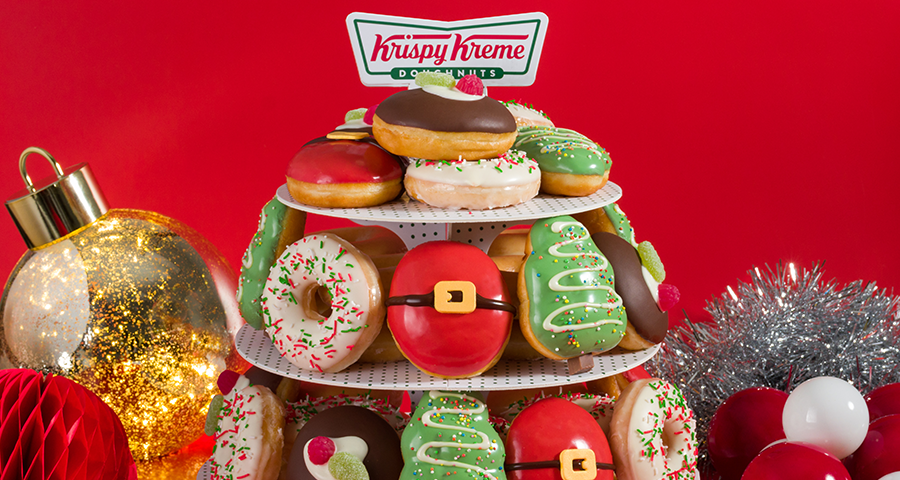 Krispy Kreme Christmas Party Pack featuring limited edition christmas doughnuts on a doughnut cake stand with christmas decorations on a red backdrop