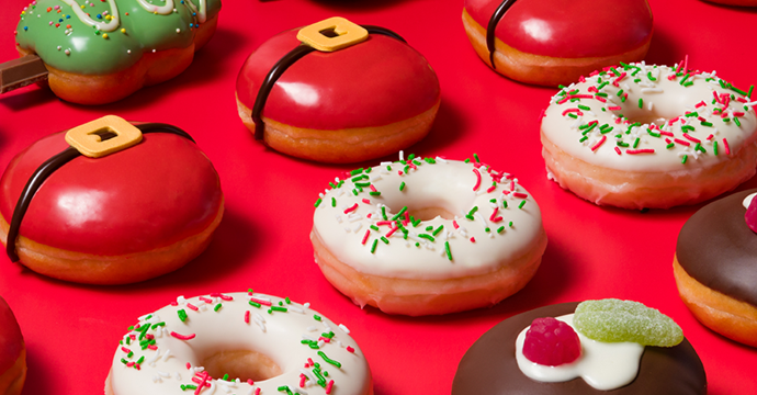 Krispy Kreme Christmas Doughnuts layed out on a red backdrop - featuring the Festive Sprinkles Ring Doughnut, the Santa Belly Filled Doughnut, the Christmas Pudding Doughnut and the Christmas Tree Shaped Doughnut