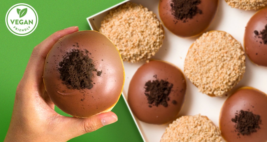Krispy Kreme Vegan Friendly Doughnuts - Green background, mixed dozen flatlay and a hand holding the Fudge Brownie Bliss Vegan Friendly Doughnut - Apple Custard Crumble Vegan Friendly Doughnut can be seen in the background also. Vegan Friendly Logo 