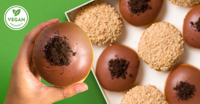 Krispy Kreme Vegan Friendly Doughnuts - Green background, mixed dozen flatlay and a hand holding the Fudge Brownie Bliss Vegan Friendly Doughnut - Apple Custard Crumble Vegan Friendly Doughnut can be seen in the background also. Vegan Friendly Logo 