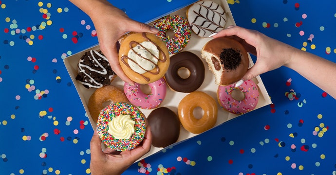 Krispy Kreme Limited Time Party Favourites Range Image of the party favourites dozen on a blue backdrop with a hand in frame holding one of the doughnuts in the range